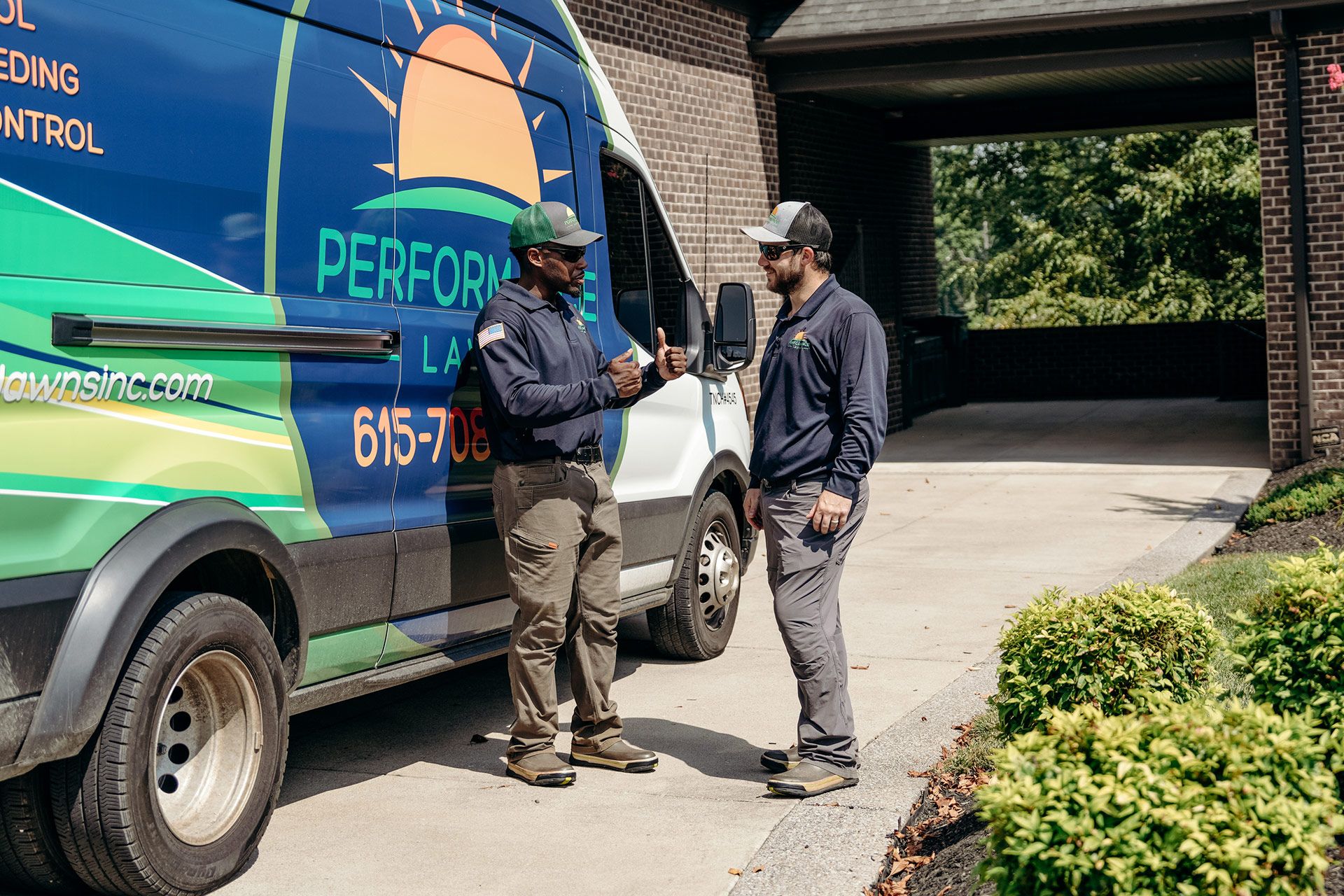 Two men in work uniforms near a van with landscaping business logo; talking outside a building.