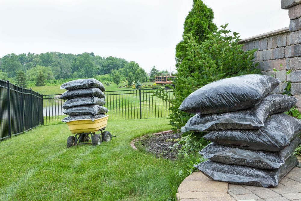 Bags of mulch stacked on a yellow wagon and near a brick wall on a green lawn with trees and a black fence.