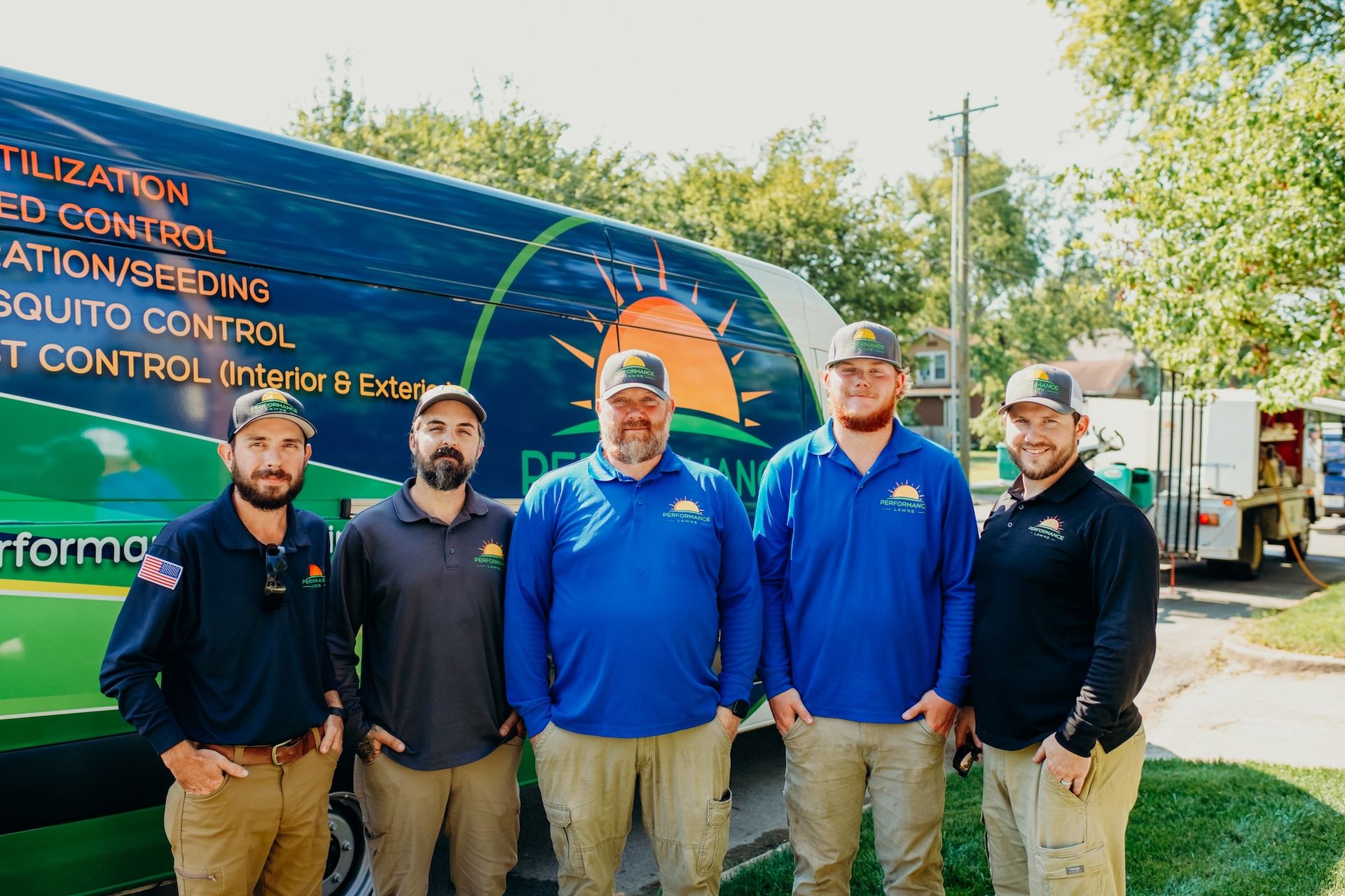 Five men in uniform pose in front of a green truck, a sunny day.
