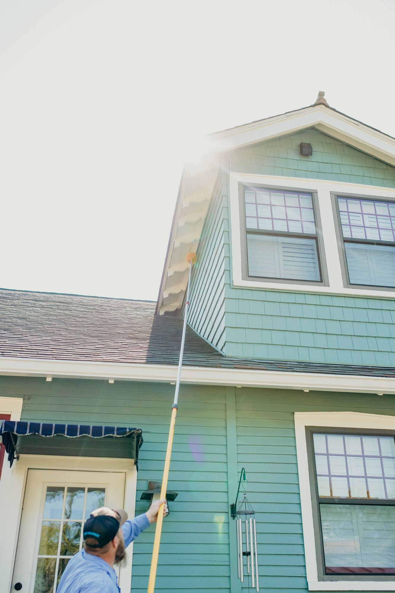 Man using pole to clean a two-story teal house. Sunny, outdoor setting.
