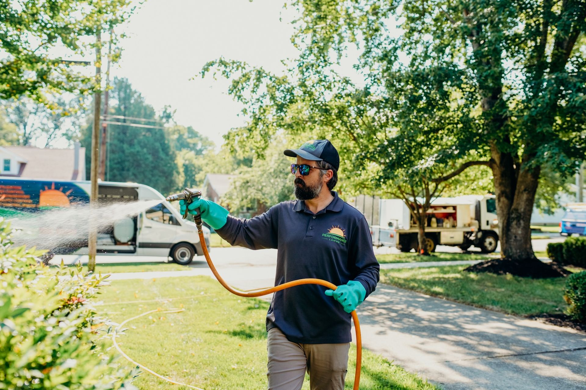 Man sprays plants with a hose in a sunny yard. He wears gloves, a cap, and a company shirt.