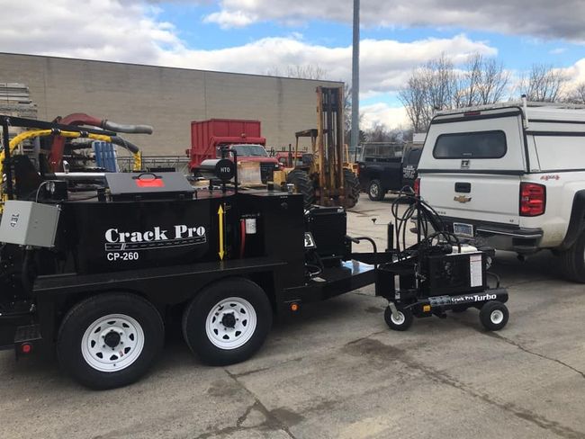 Black Crack Pro road crack sealing trailer with attached applicator, white wheels, parked near truck.