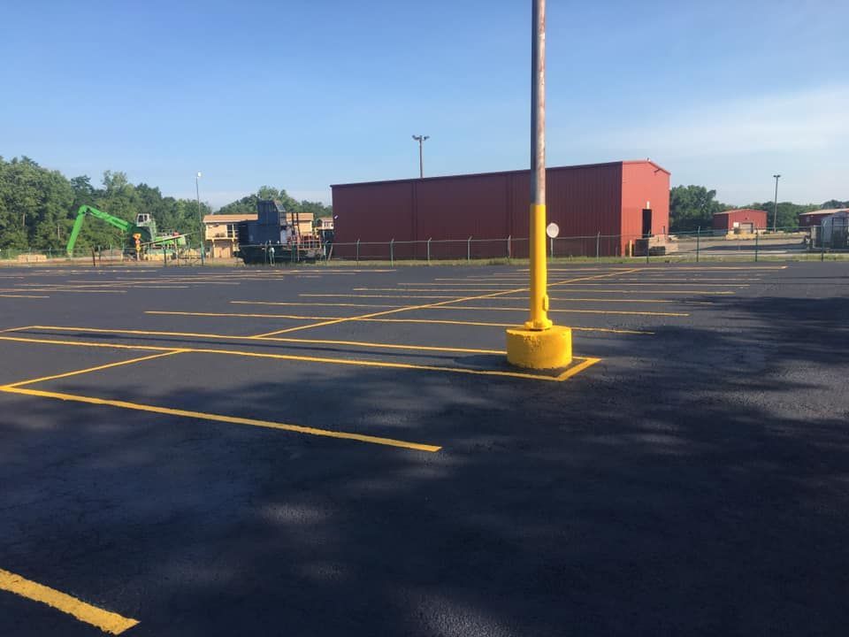 Newly paved parking lot with yellow lines, a red building in the background, and a yellow-painted light pole base.