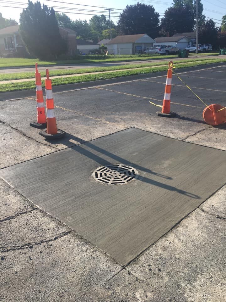 Freshly poured concrete square with drain, marked by orange cones on asphalt.