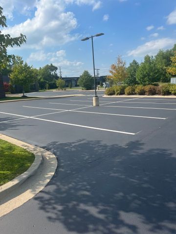 Newly paved parking lot with yellow lines, in front of a brick building and trees under a blue sky.