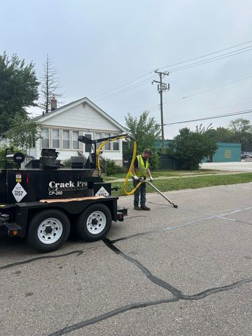 Person applying sealant to a road crack, using a metal applicator. Includes an orange traffic cone.