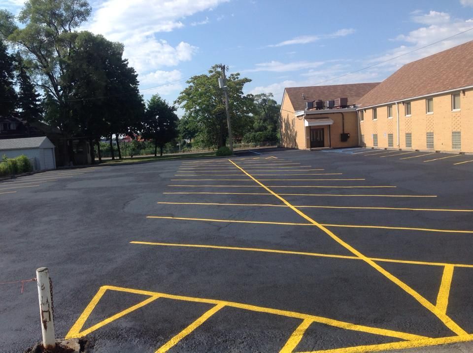 Newly paved, empty parking lot with yellow painted lines in front of a brick building under a sunny sky.