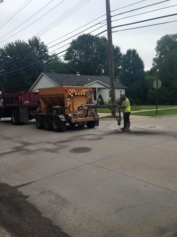 Worker operating asphalt patching equipment on a street, with a truck, house, and utility pole in the background.