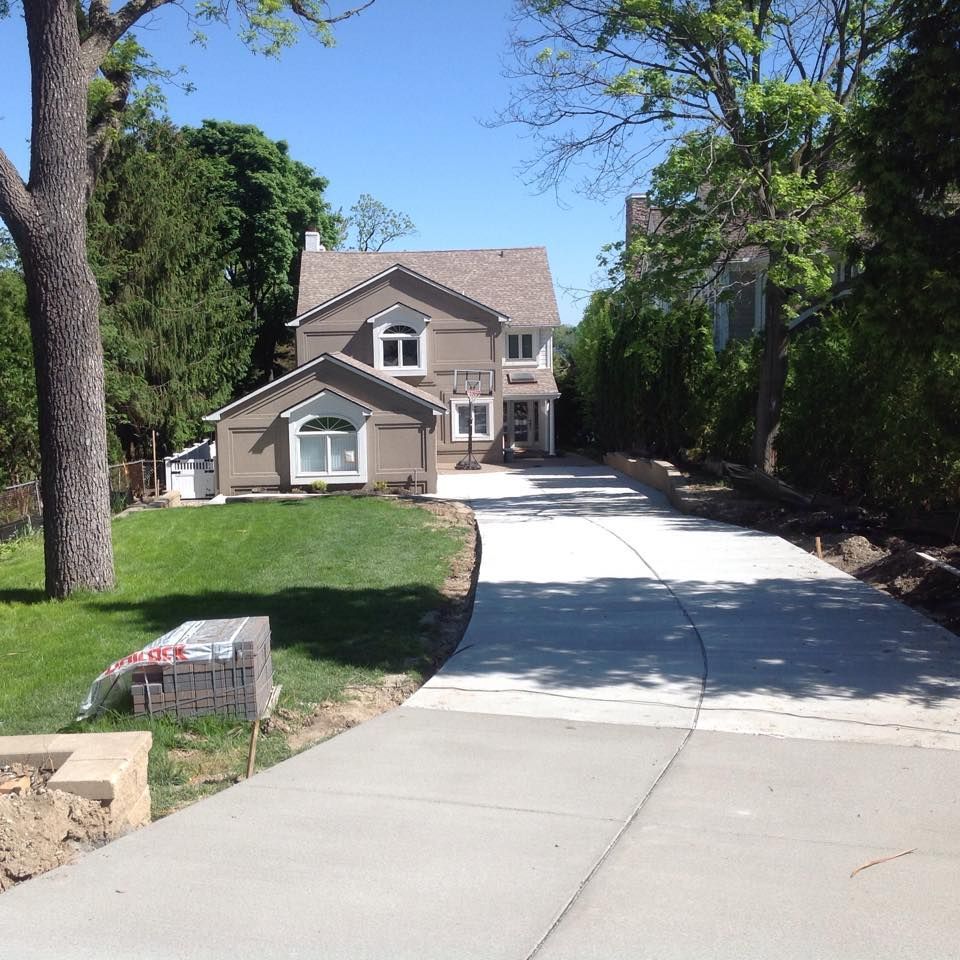 New concrete driveway leading to a two-story house with a brown roof and a green lawn on a sunny day.