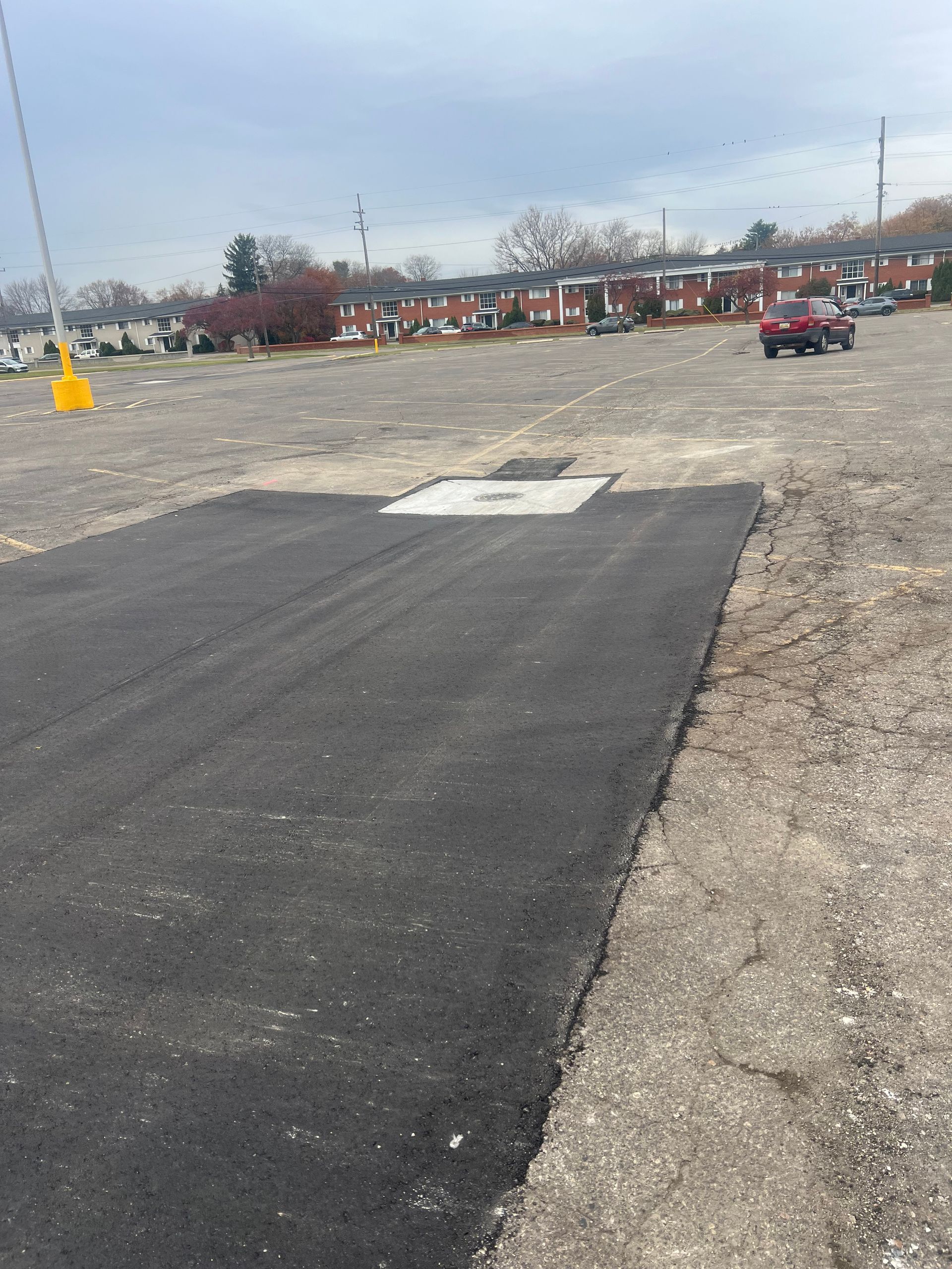 Fresh black asphalt patch in a gravel parking lot with a small white utility cover nearby.