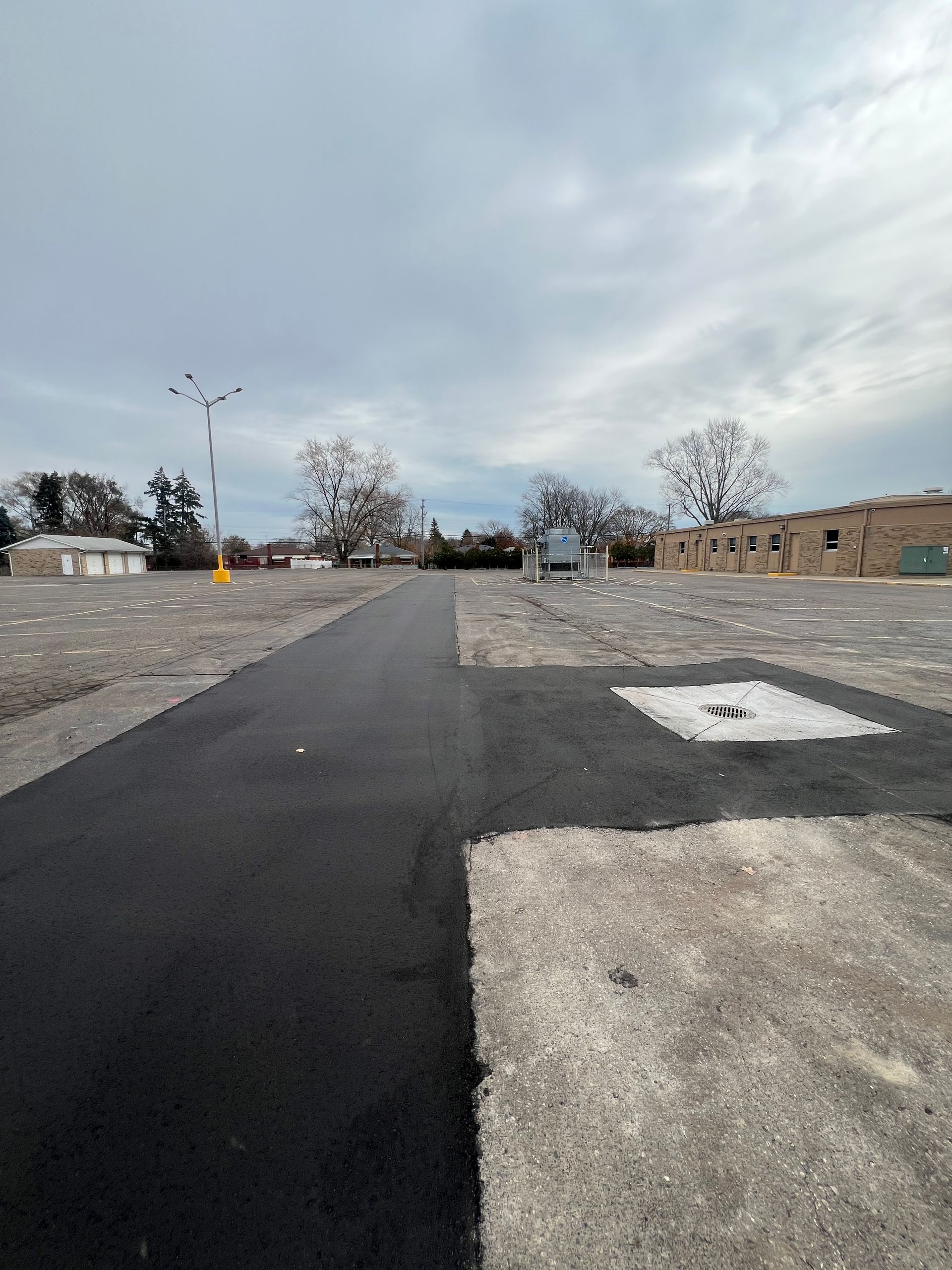 Empty paved parking lot under a cloudy sky, with a road on the left and buildings in the distance