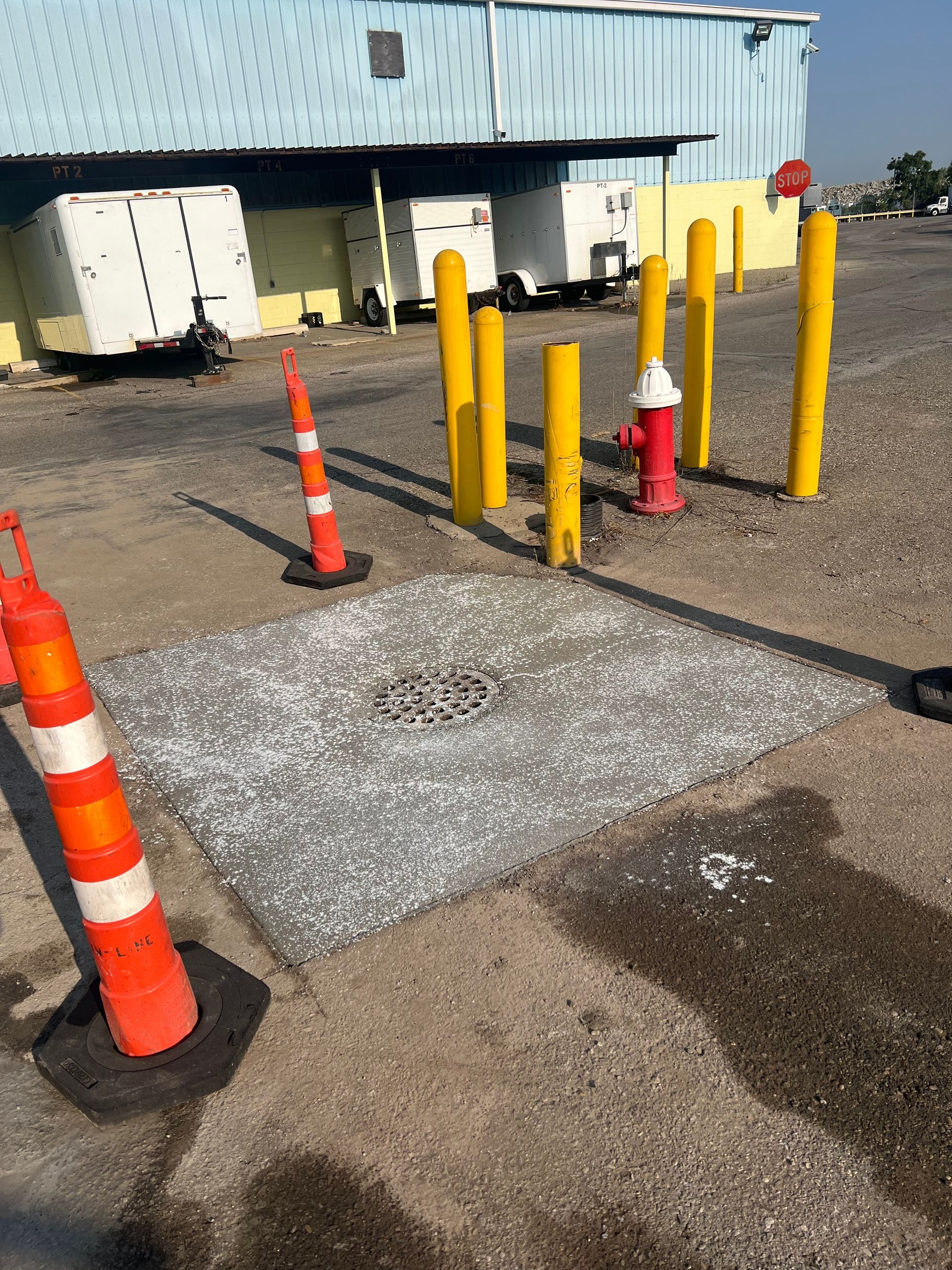 Orange traffic cones and yellow bollards on a gravel lot beside a building and utility tank under a metal awning