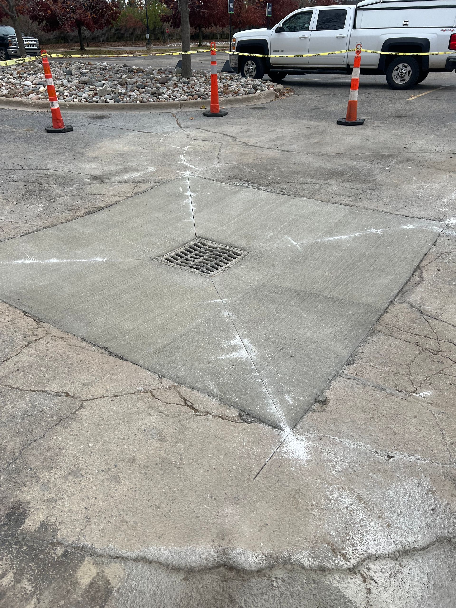 Cracked concrete parking lot with a square drain and orange cones near a white pickup truck.