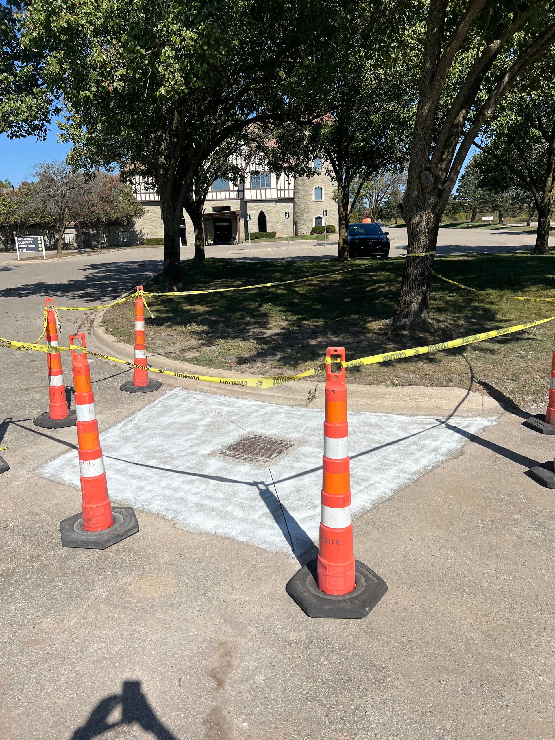 Orange cones and caution tape block off a damaged sidewalk area near trees and a building.