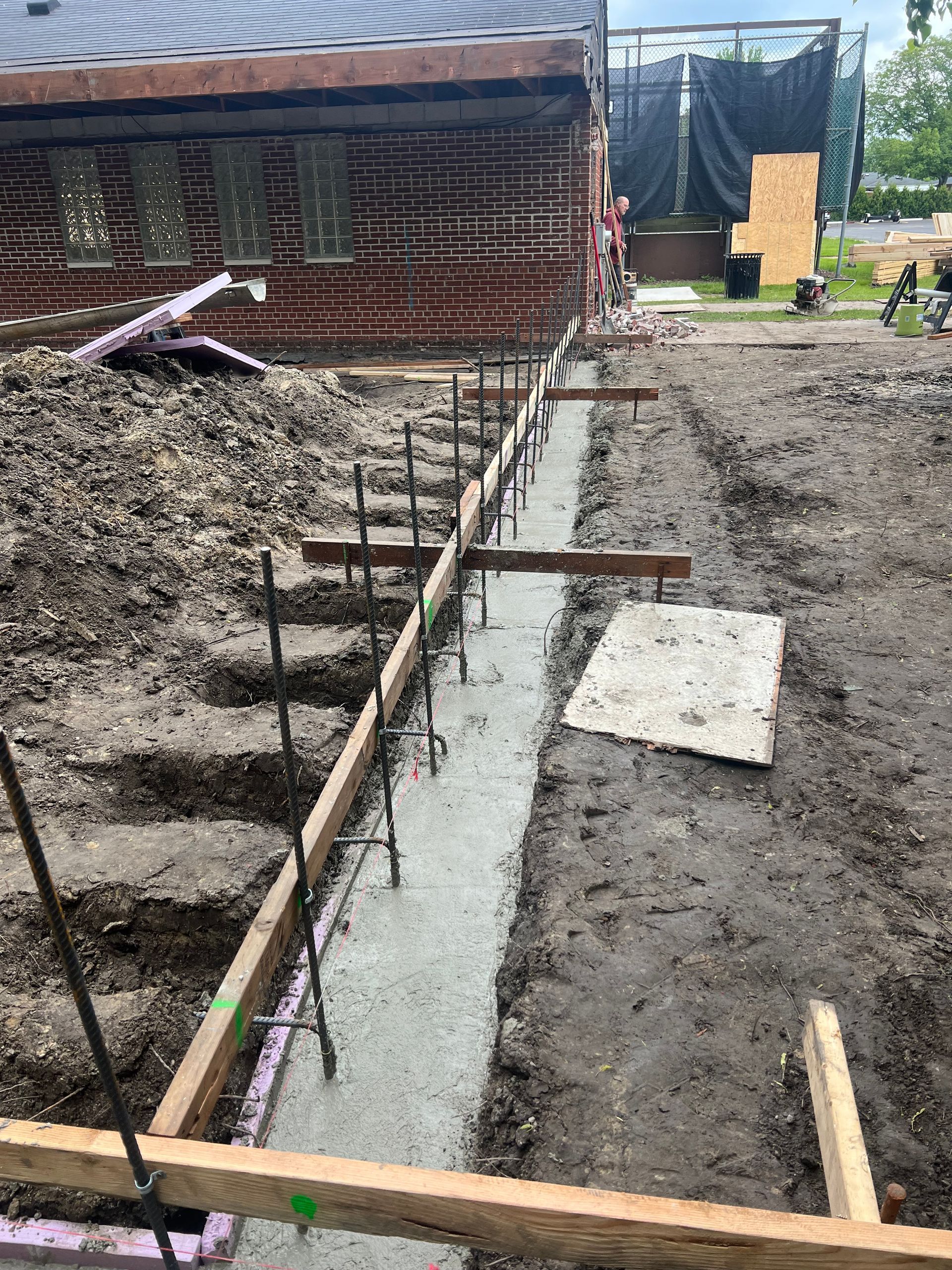 Construction trench with wooden formwork and concrete beside a brick building.