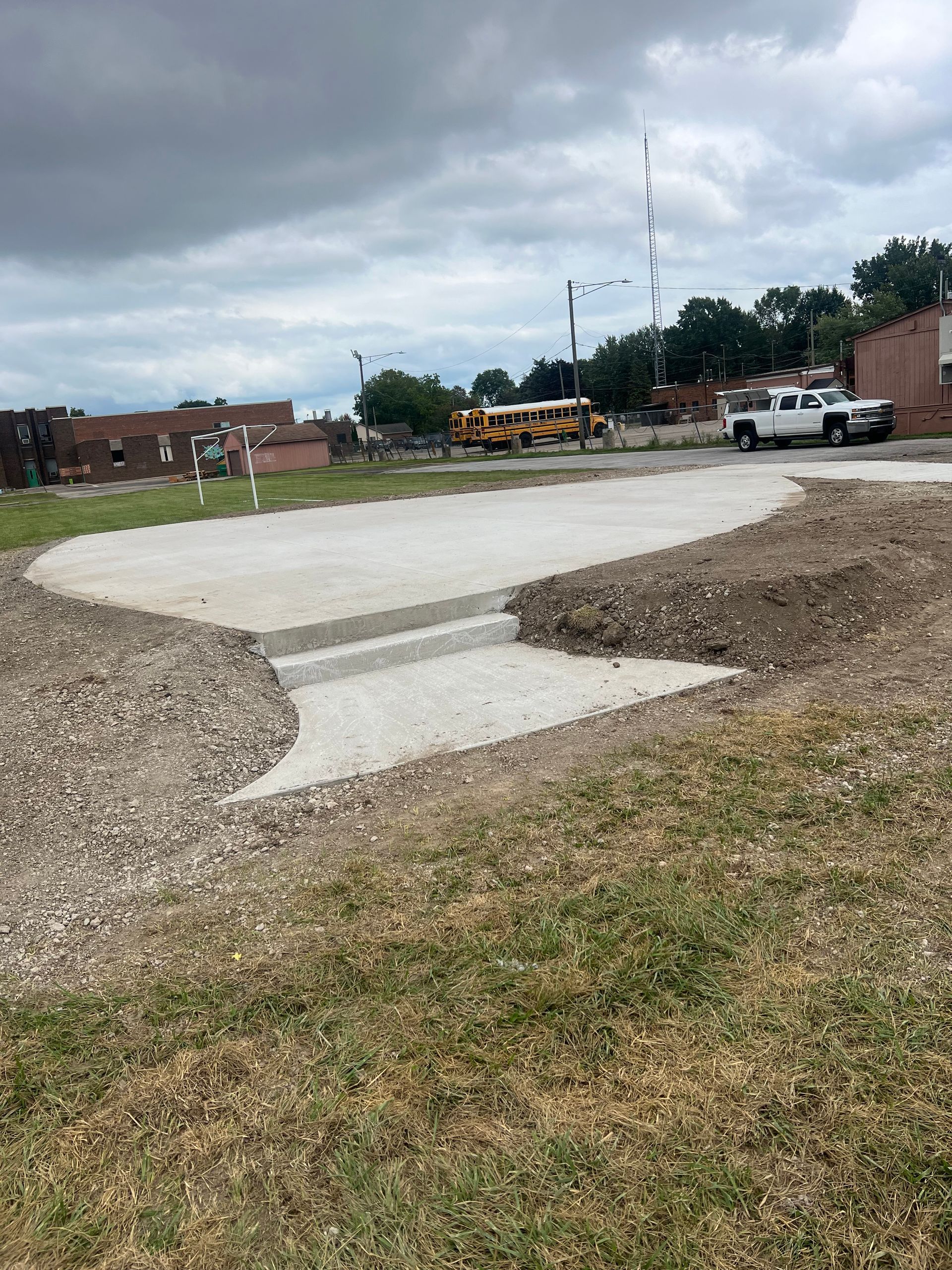 Concrete path in a grassy lot with parked vehicles and equipment under a cloudy sky