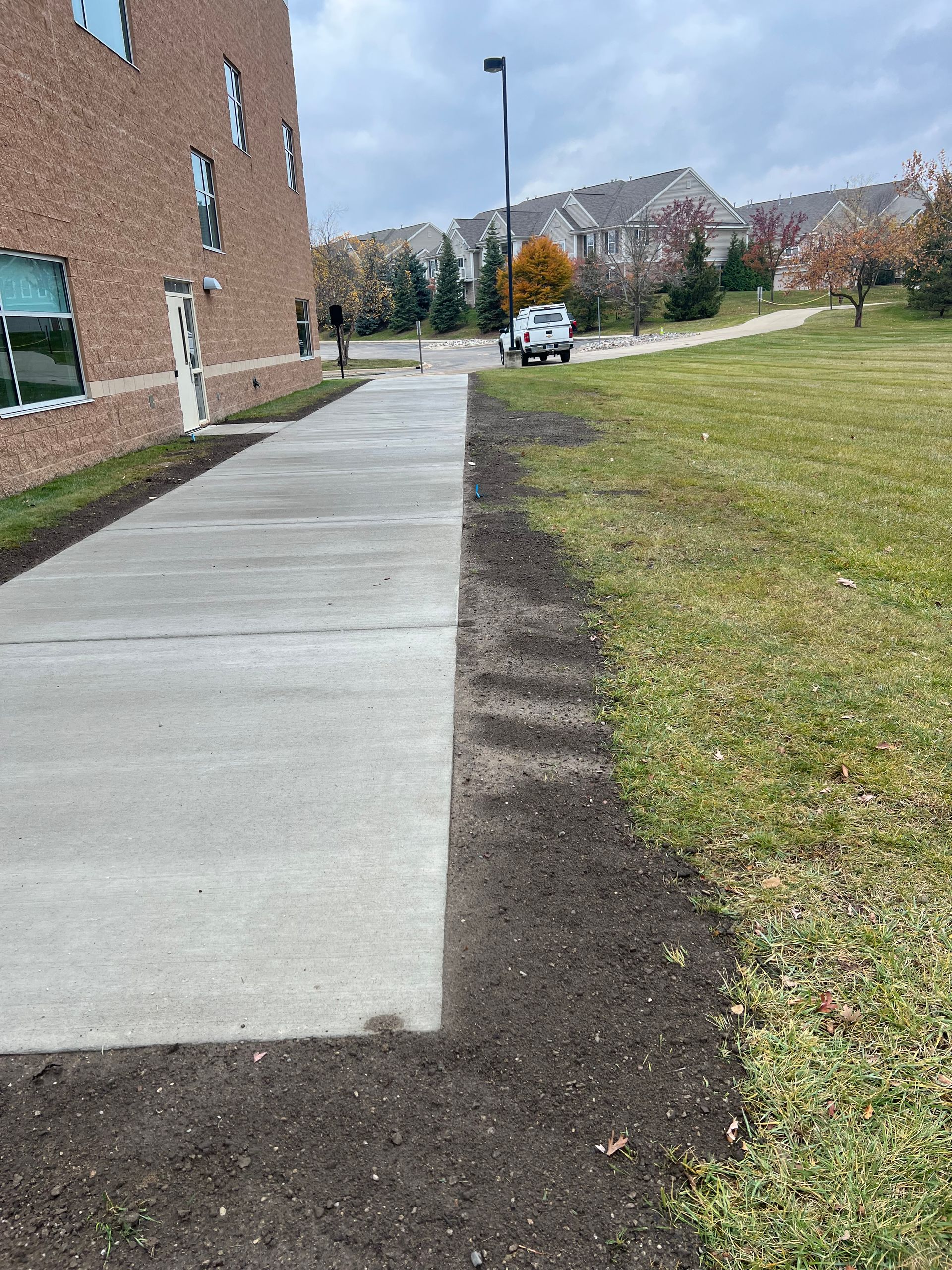 Concrete sidewalk beside a brick building, bordered by grass and a parked car on an overcast day