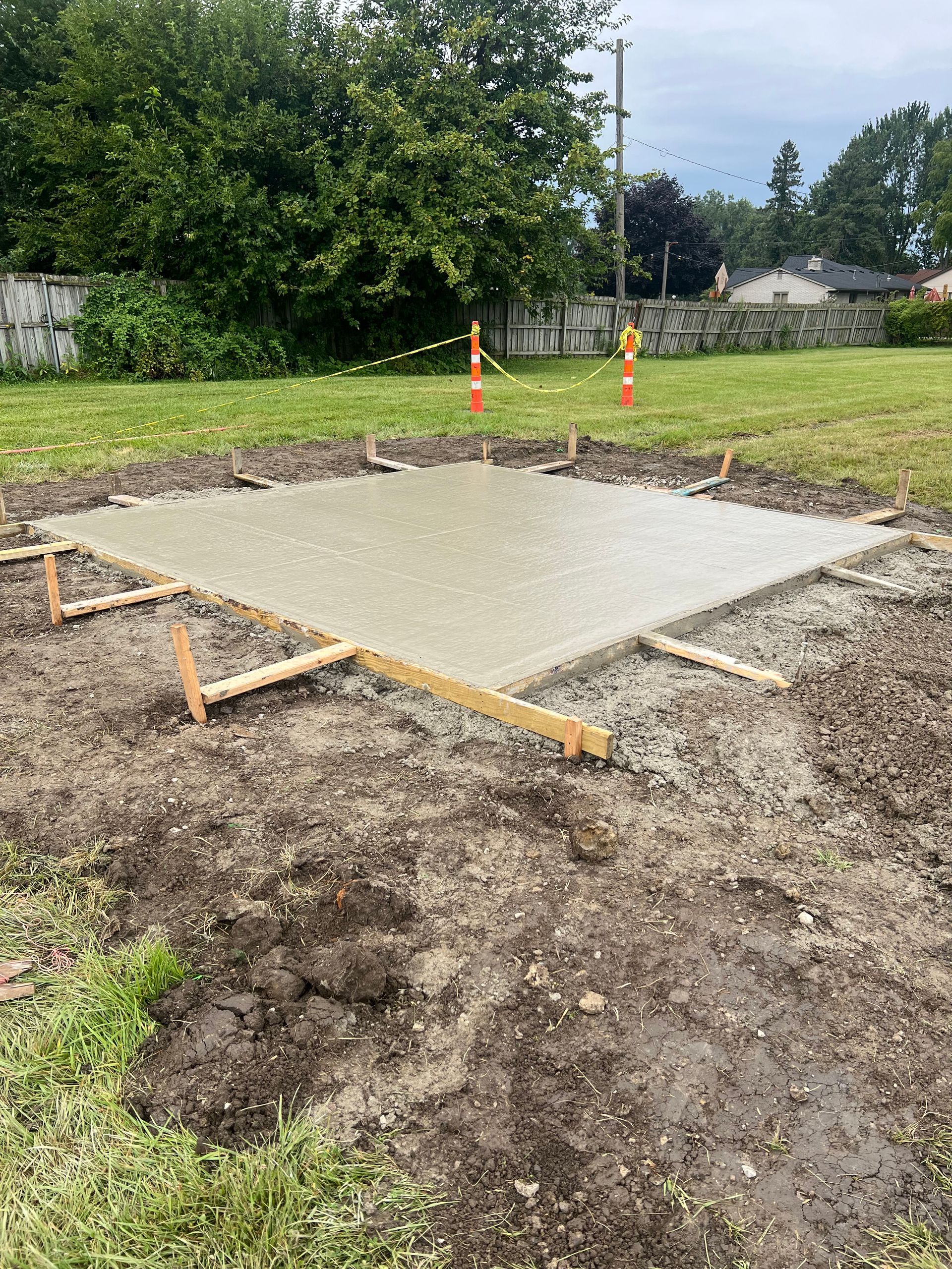 Fresh concrete slab in a yard, bordered by dirt and wooden forms, with orange cones and trees in the background