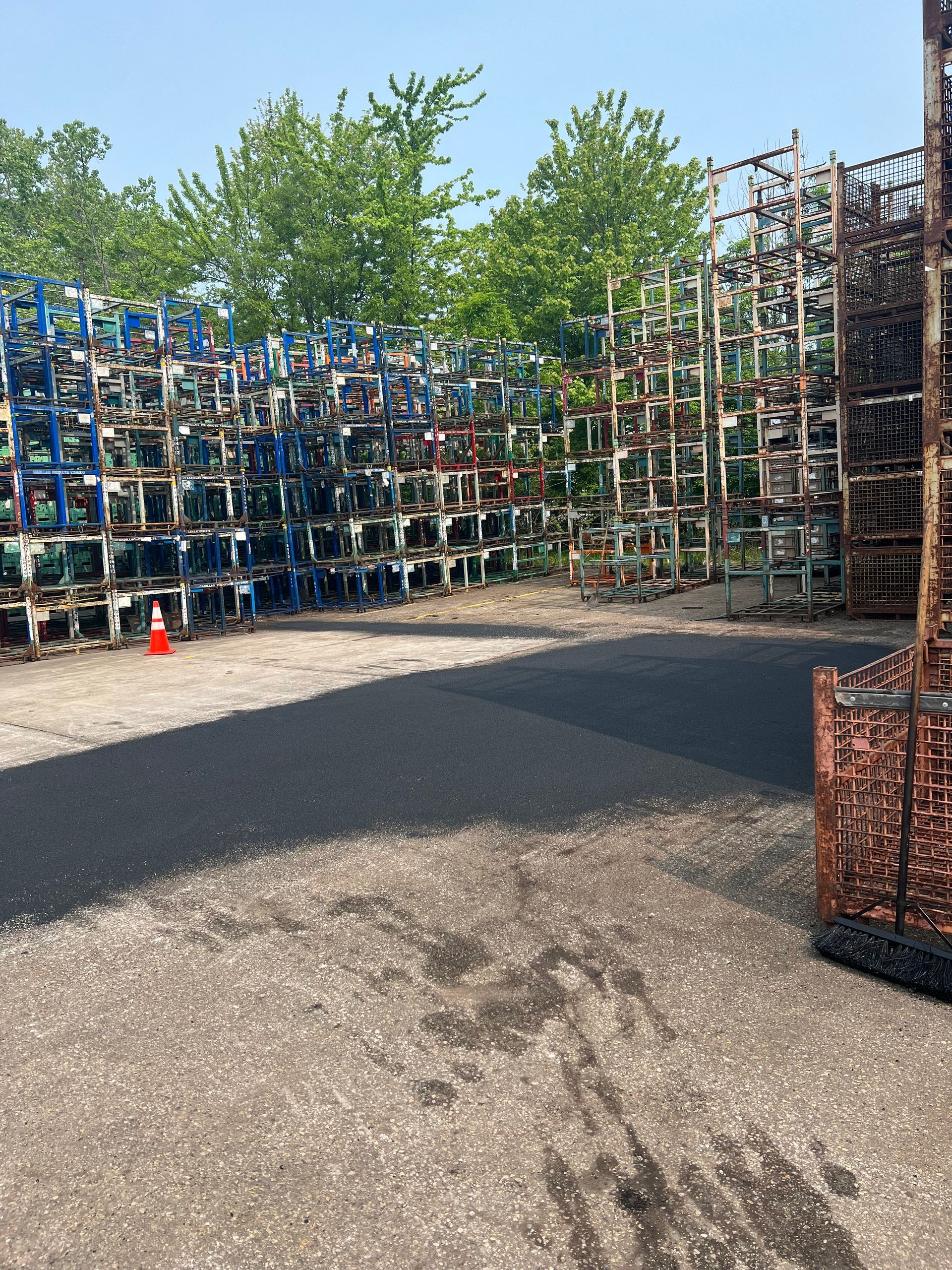 Outdoor yard with stacked metal cages and bins beside gravel ground and green trees under a clear sky