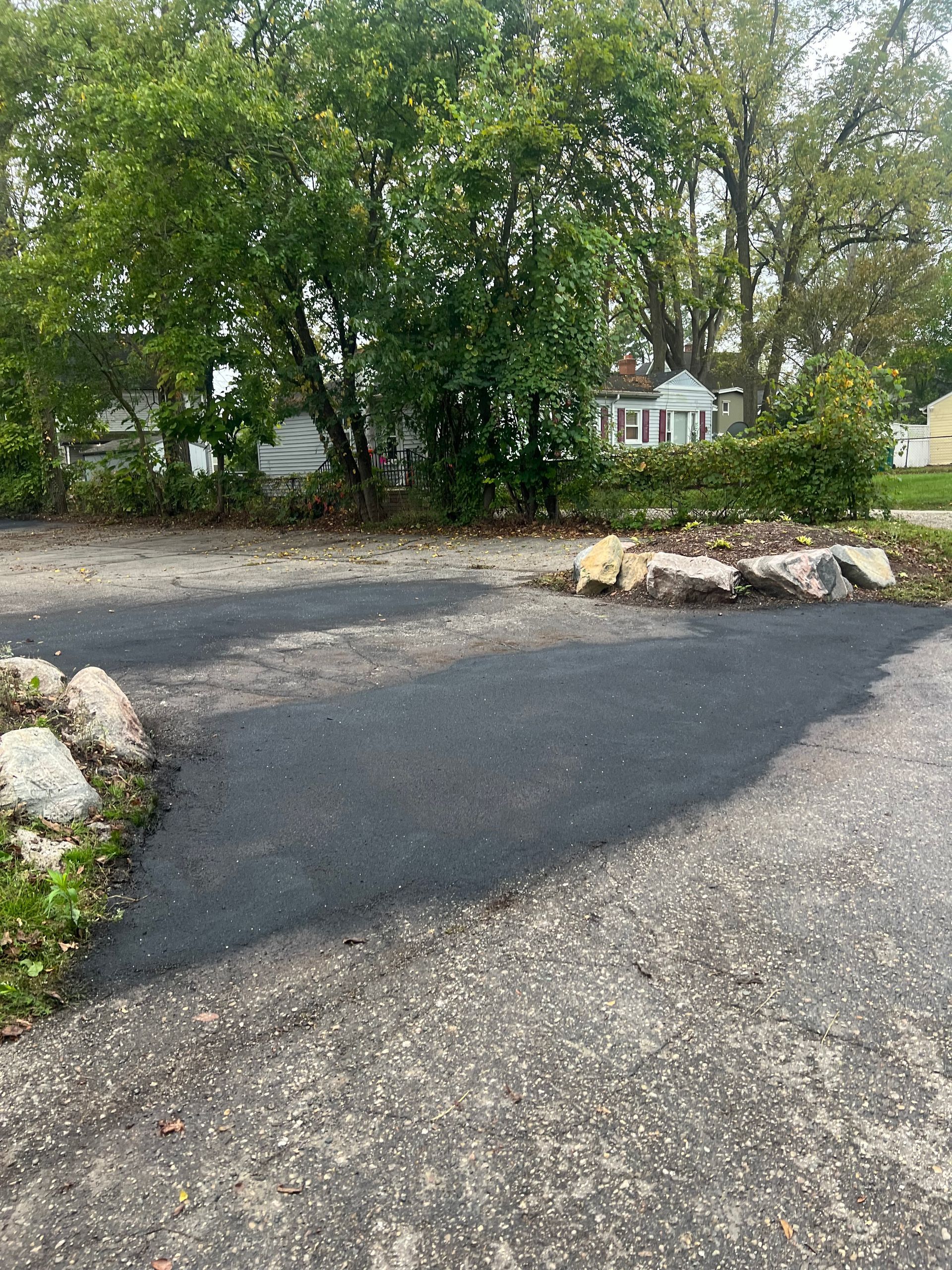 Gravel driveway beside trees and rocks, with a house partially visible in the background.