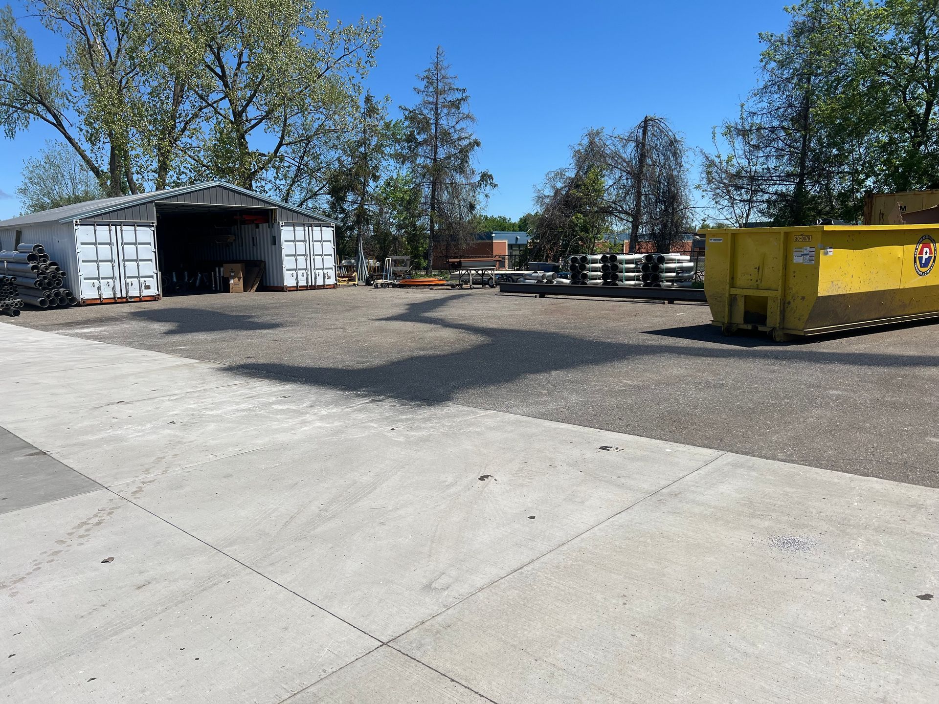 Empty paved lot beside a white garage and yellow dumpster under a clear blue sky