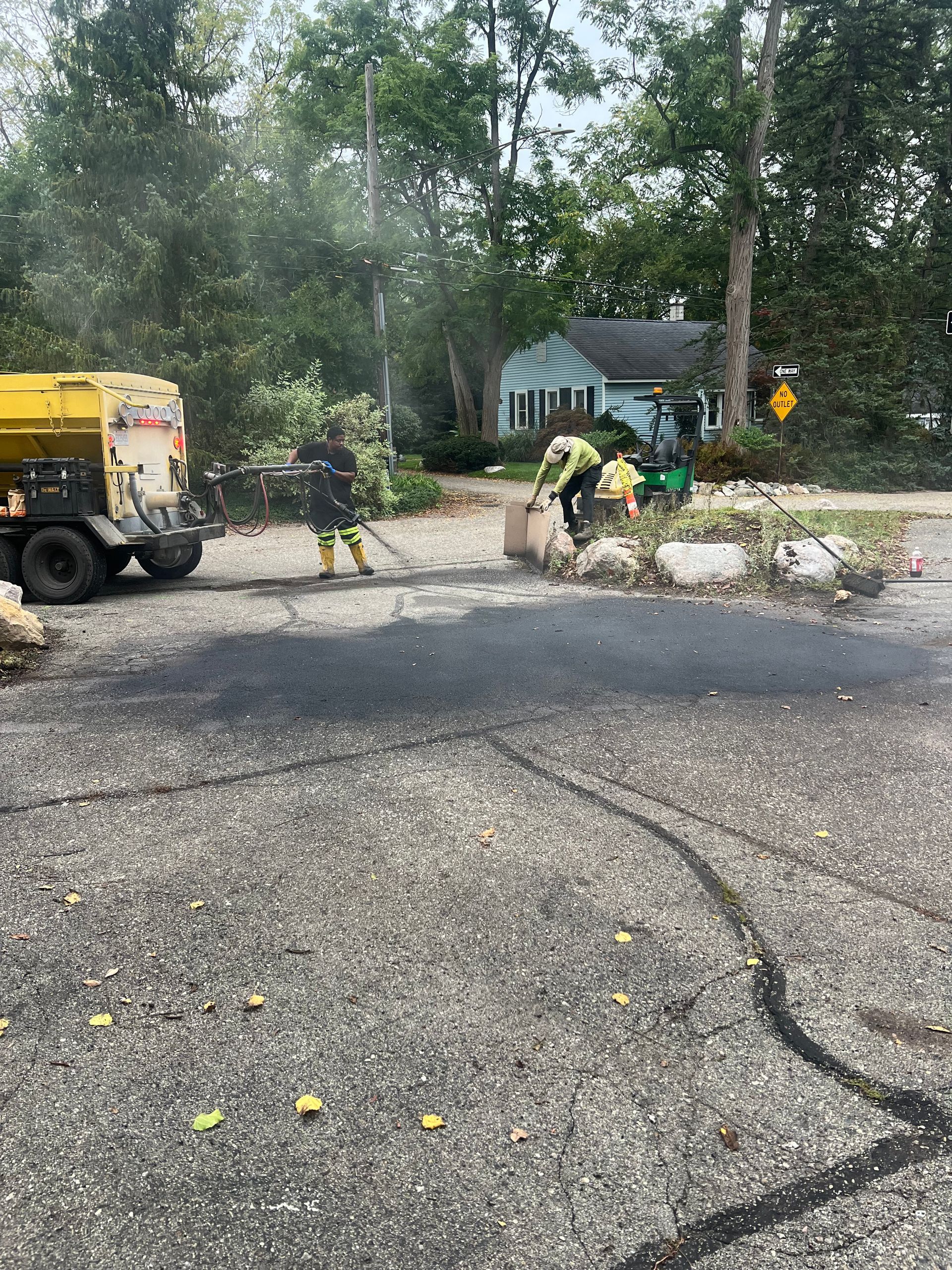 Roadwork crew repairing a cracked street beside a tree-lined home, with a yellow truck and workers clearing debris