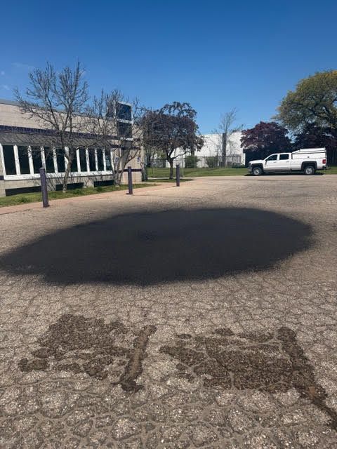 Cracked parking lot with a large dark stain, trees, building on left, and a white truck in the background