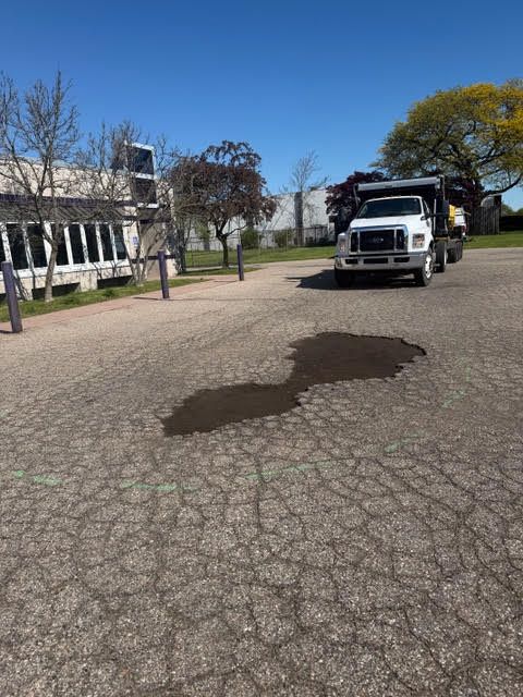 White truck parked on a cracked street near a building, with a puddle in the foreground and trees under a blue sky.