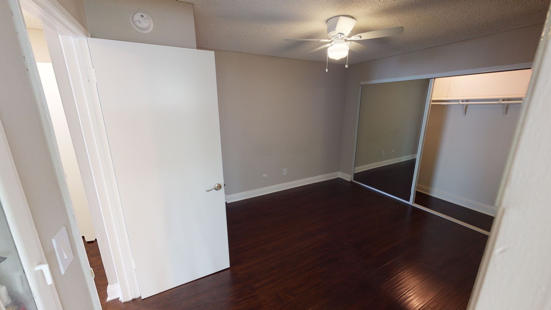Empty bedroom with dark hardwood floors, gray walls, and a mirrored closet.