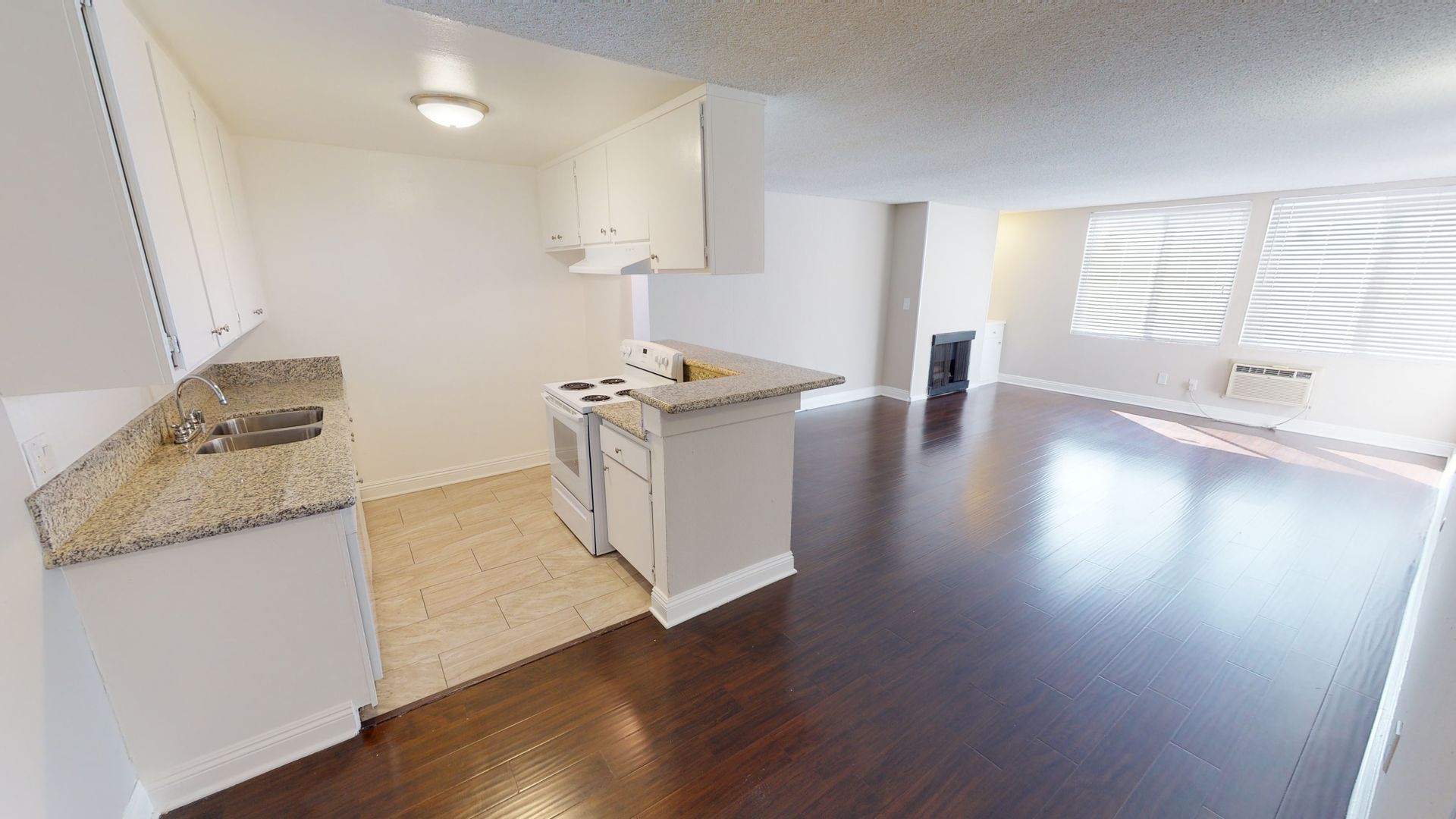 Kitchen and living area of a small apartment with white cabinets, dark wood floors, and a window.