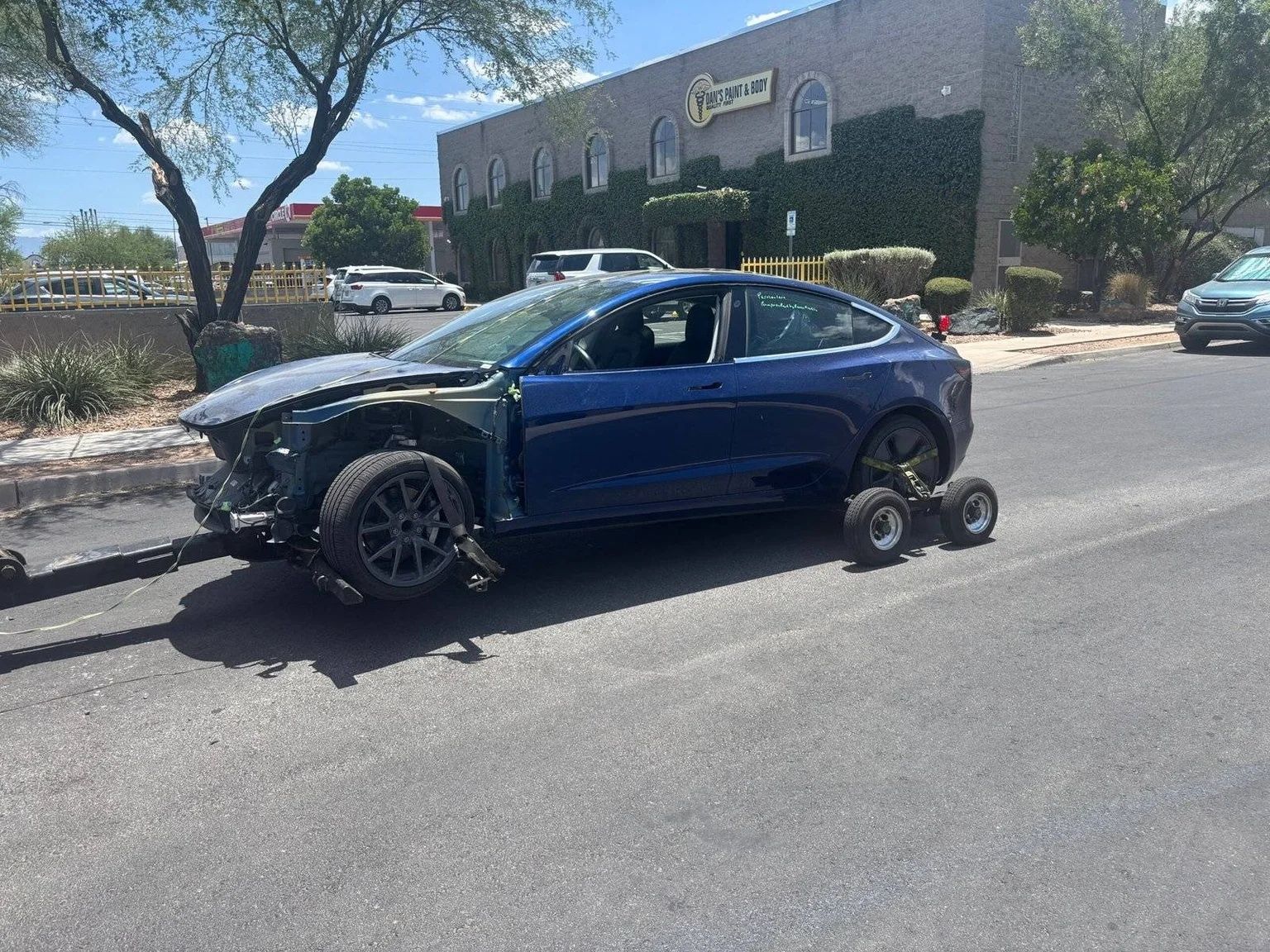 Blue car with front-end damage being towed on a road outside a storefront