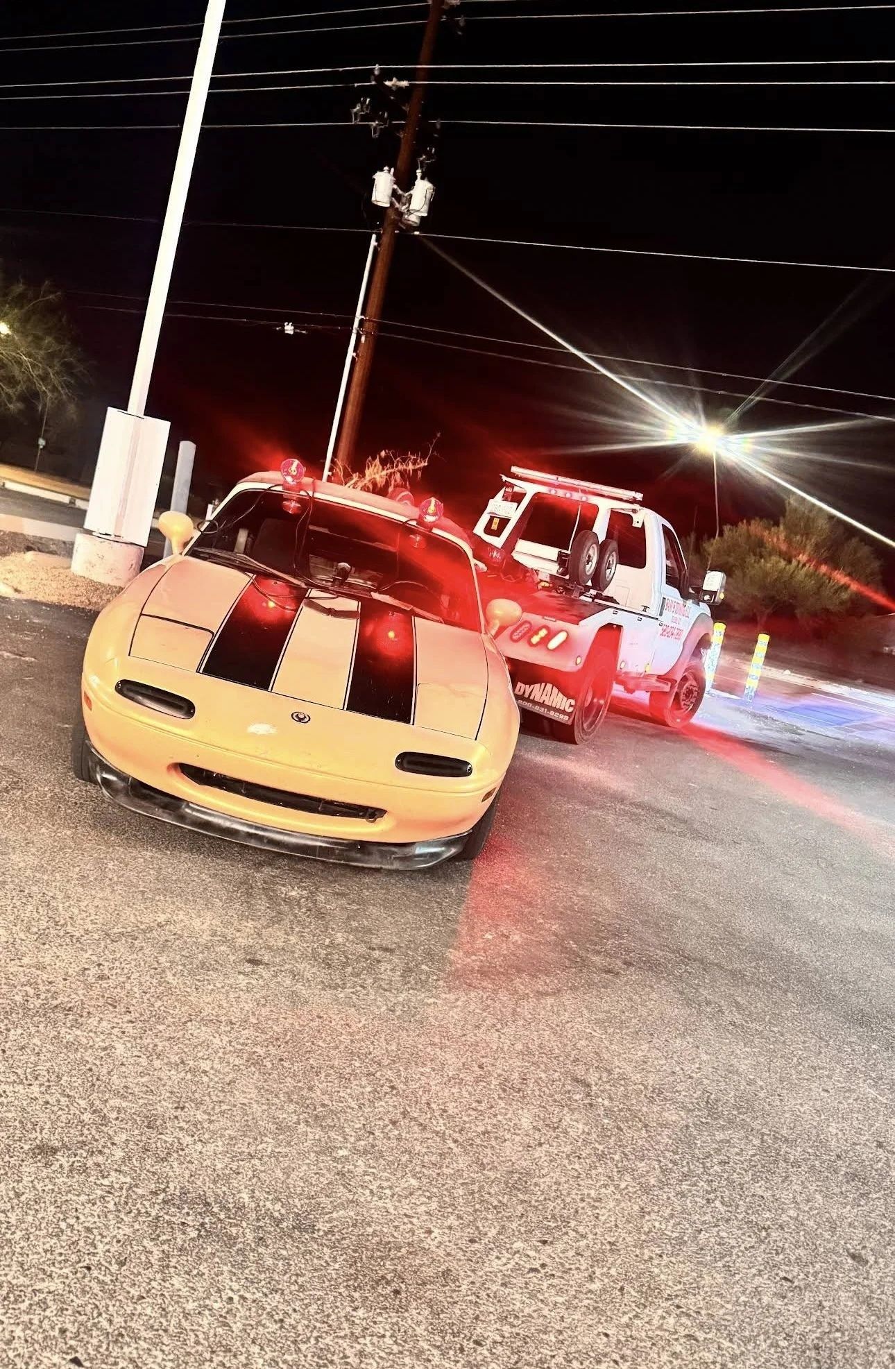 Yellow sports car with black racing stripes on a street at night, near a tow truck with flashing lights