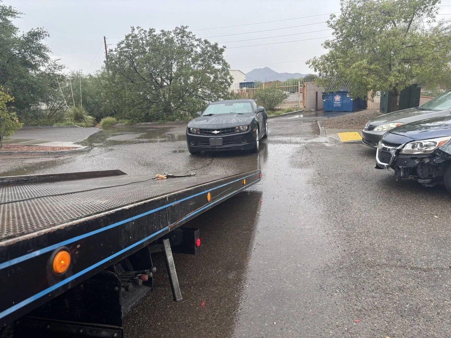 Rainy parking lot with cars beside a flatbed tow truck, viewed from the truck’s ramp