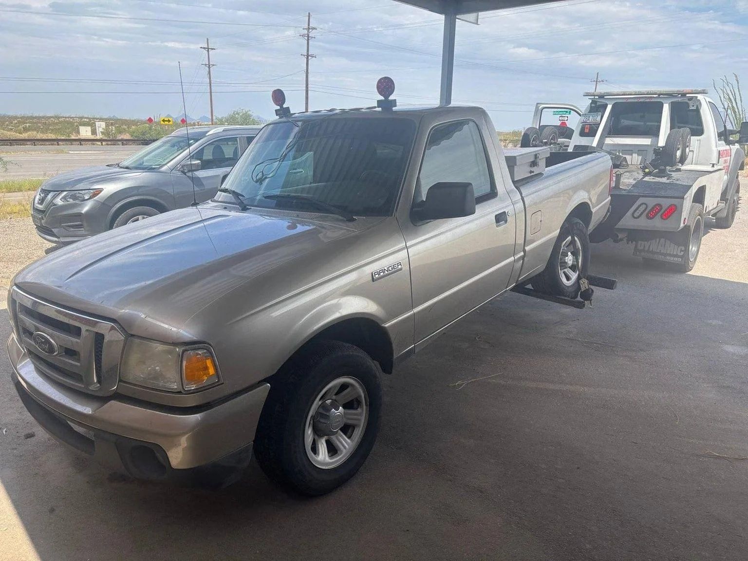 Beige pickup truck parked at a gas station beside a tow truck during roadside service