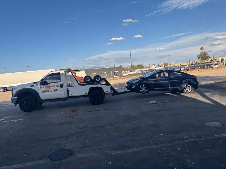 Tow truck towing a black sedan in a parking lot under a blue sky