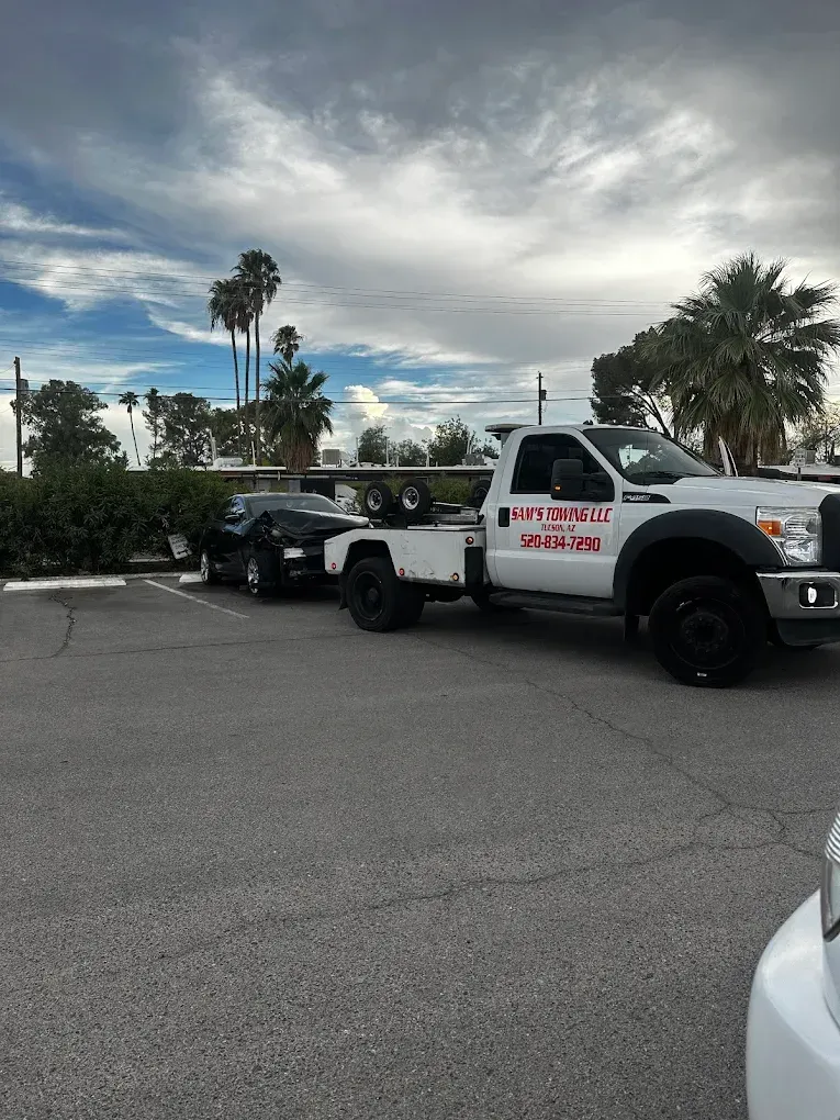 White tow truck hauling a black car in a parking lot under a cloudy sky.