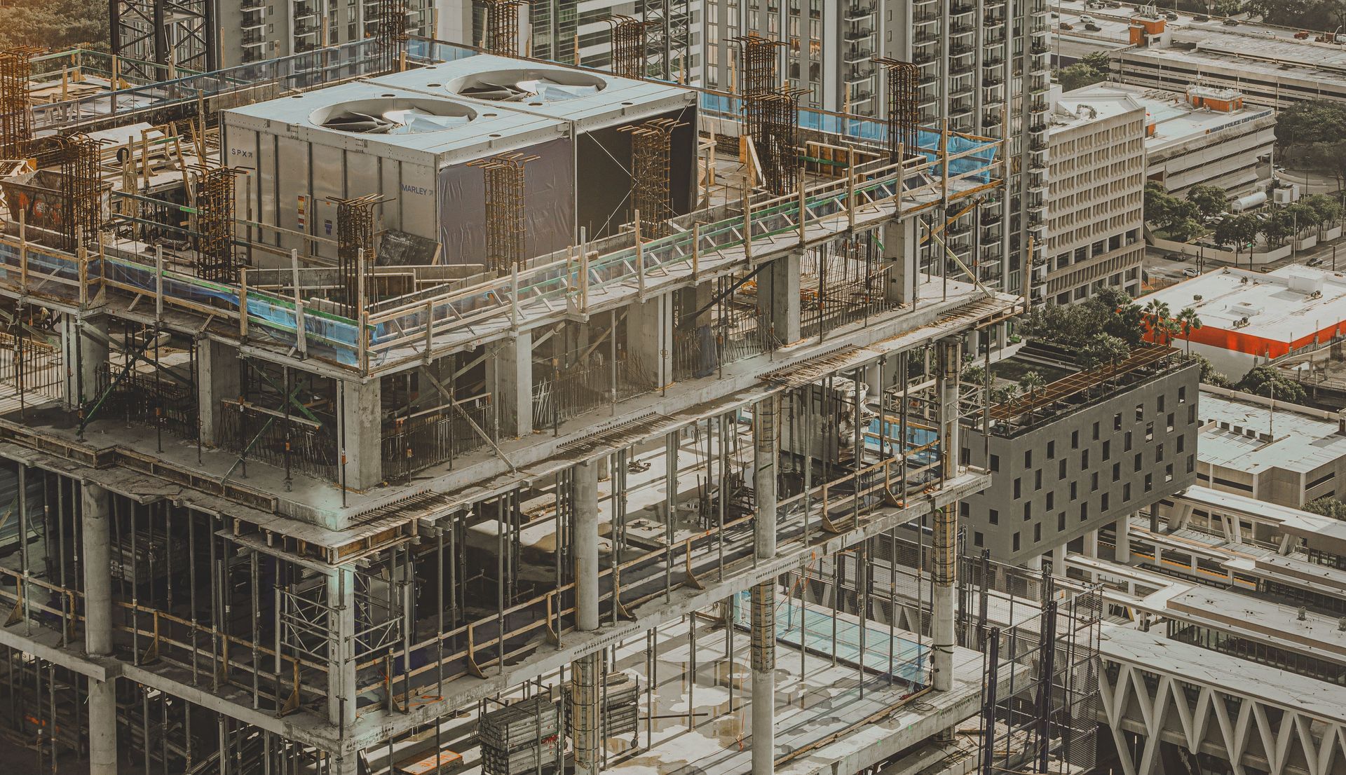 Steel-framed high-rise under construction in a dense cityscape, with cranes and scaffolding visible