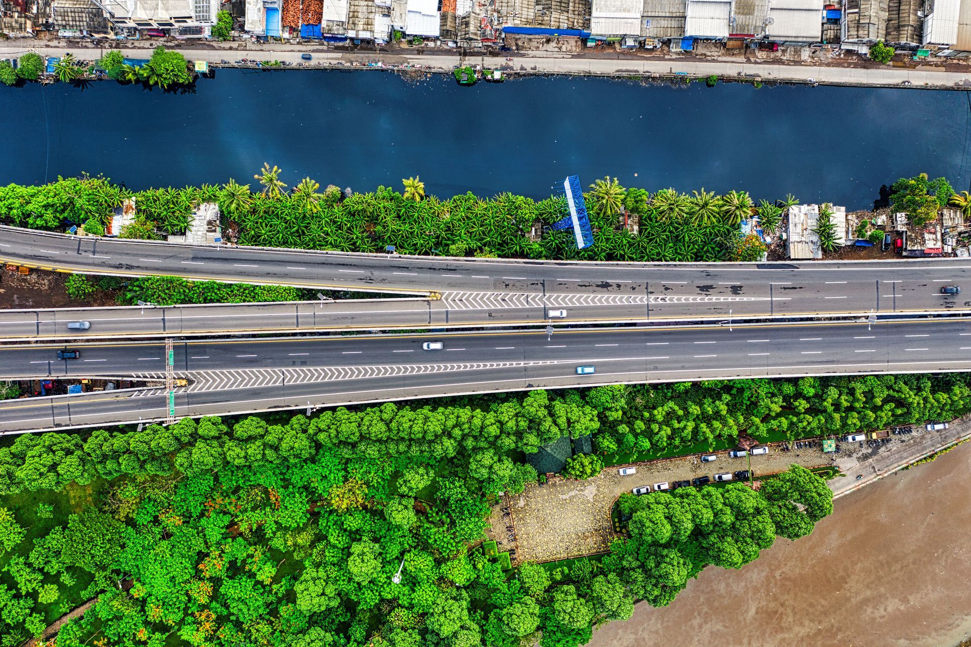 Aerial view of a road beside a blue river, with green trees and buildings along the banks.