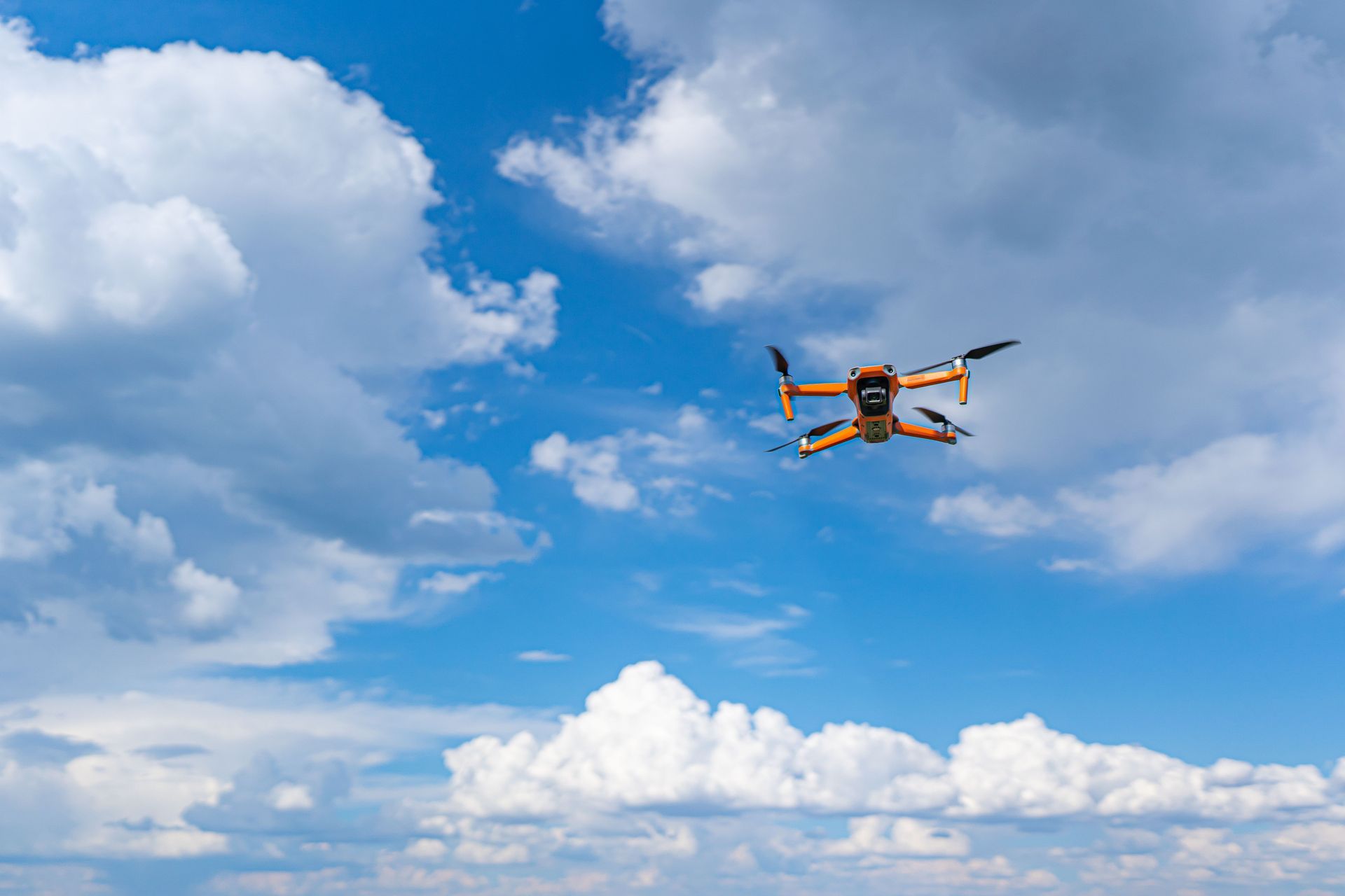 Orange drone flying in a bright blue sky with white clouds