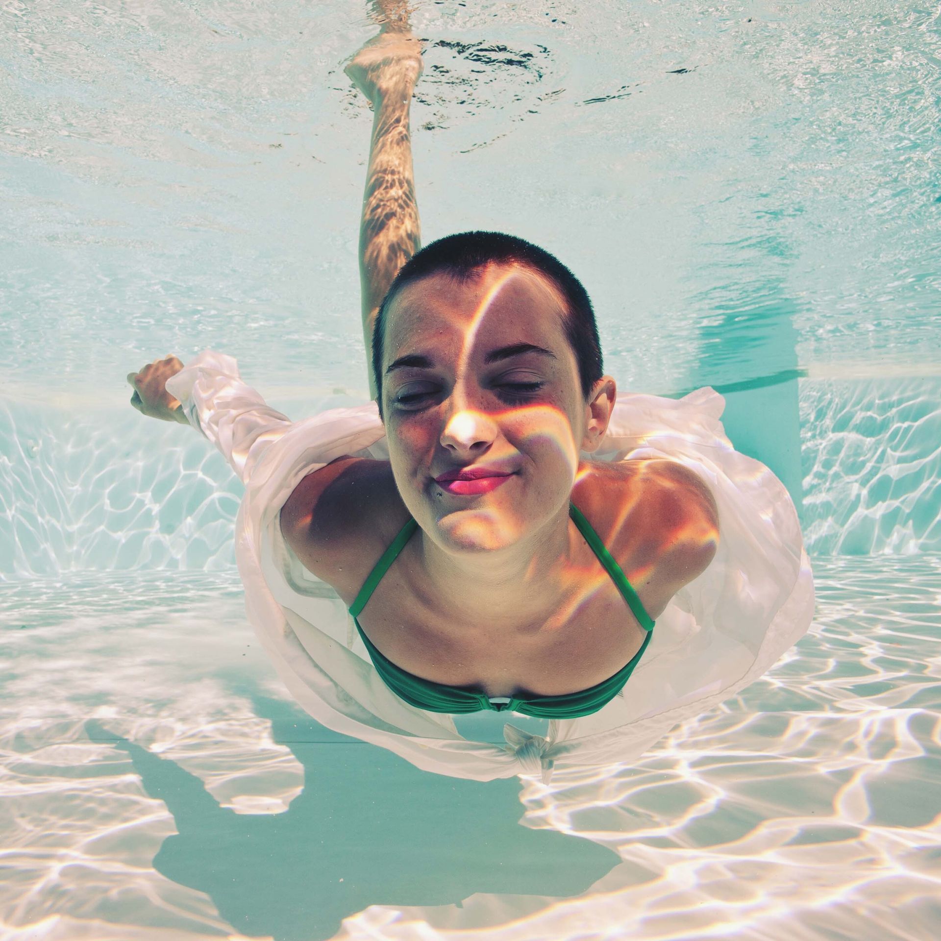 Woman with a shaved head swims underwater, wearing a white dress and green bikini top, smiling, in a pool.