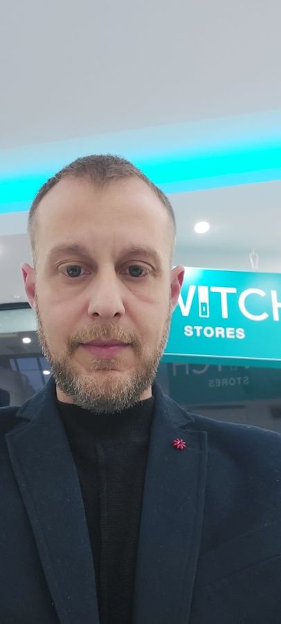 Άνδρας με σκούρο κοστούμι και μαύρο ζιβάγκο βγάζει selfie σε ένα κατάστημα με μια γαλαζοπράσινη πινακίδα πίσω του