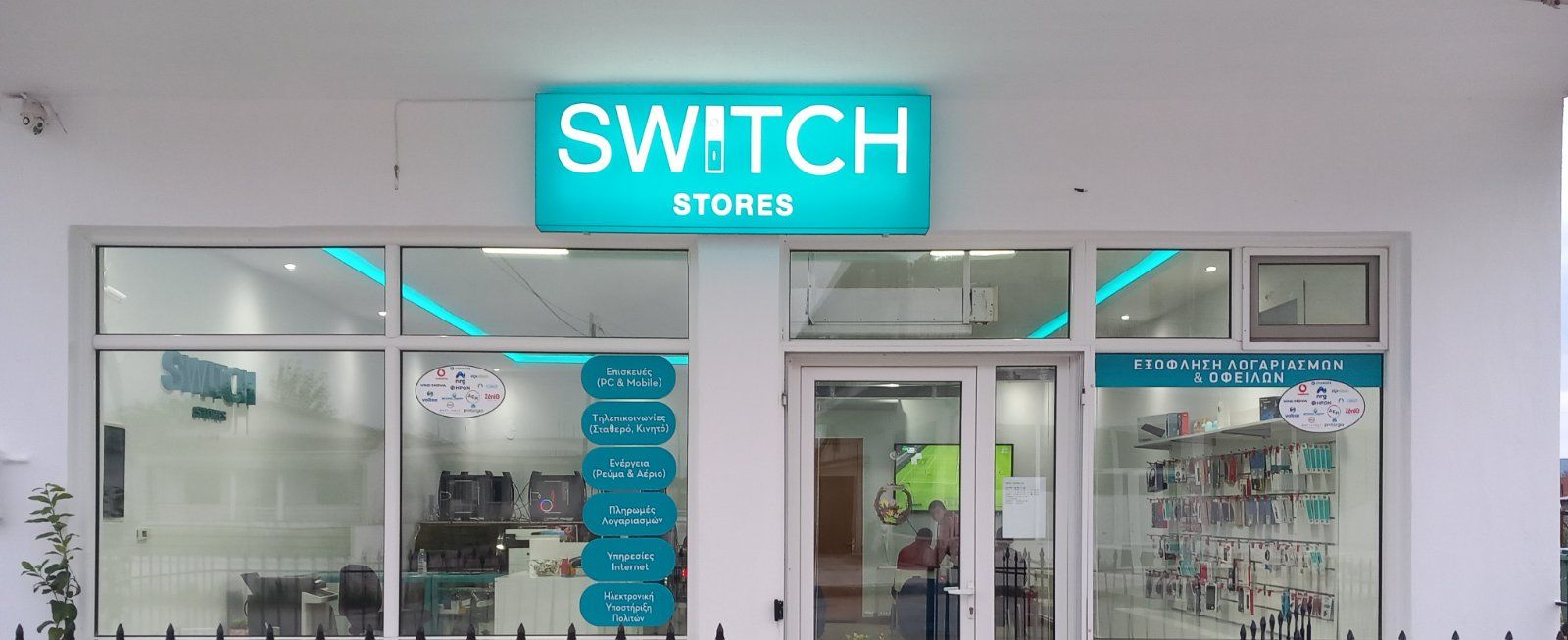Εξωτερικό των καταστημάτων Switch με τιρκουάζ πινακίδες. Διαφανή γυάλινα παράθυρα δείχνουν το εσωτερικό ενός καταστήματος με εμπορεύματα.