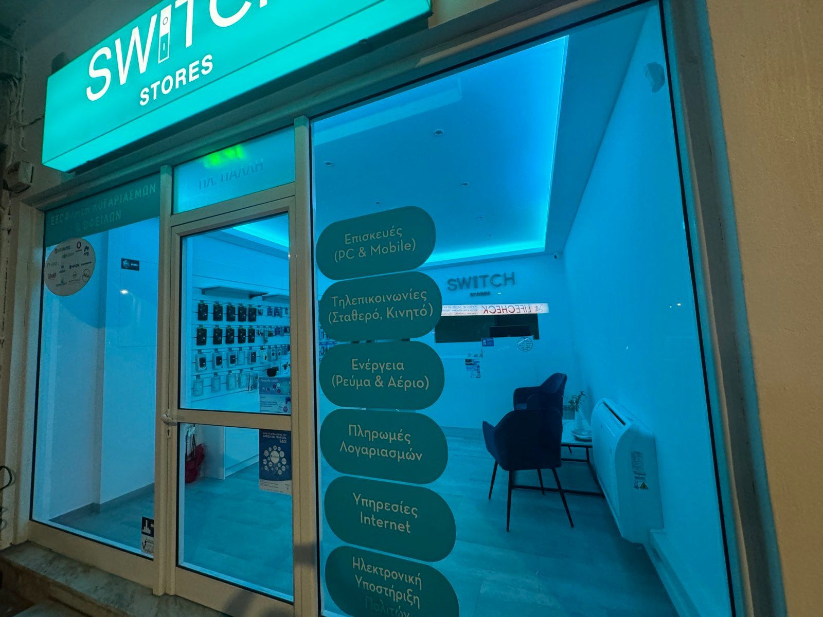 Βιτρίνα καταστήματος Switch Stores με μπλε τζάμια και φωτεινή επιγραφή. Υπηρεσίες που αναγράφονται στο τζάμι.