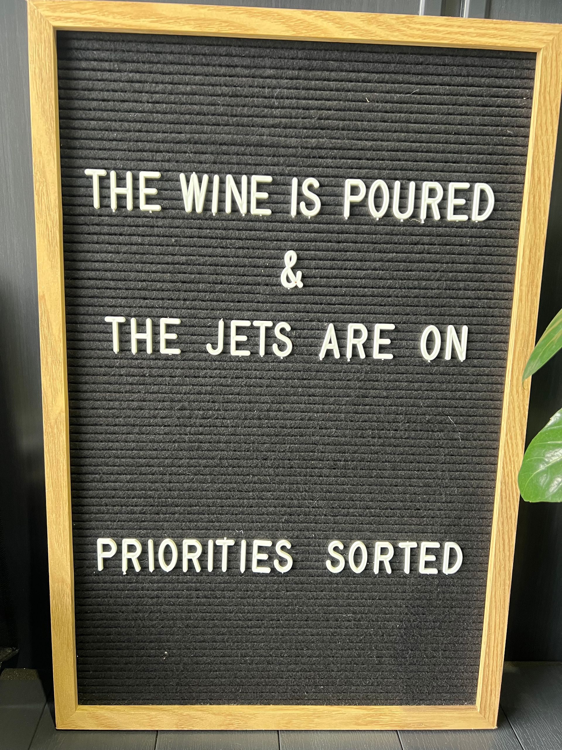 Black letter board with white text: “The wine is poured & the jets are on priorities sorted”
