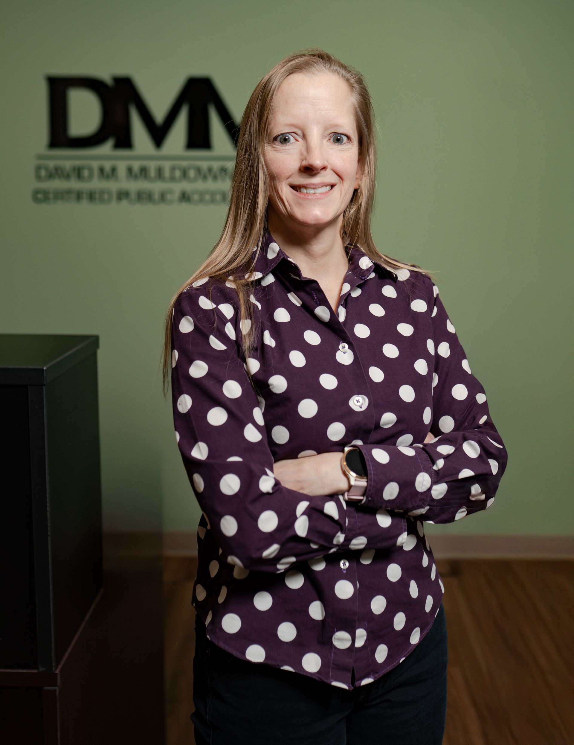 A woman in a polka dot shirt stands with her arms crossed in front of a sign that says dmn