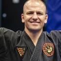 A man in a black karate uniform is smiling with his arms outstretched.