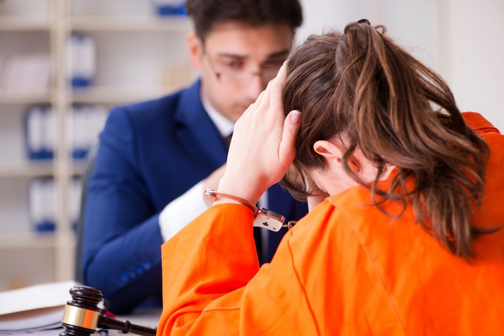 Woman in orange jumpsuit, handcuffed, with head in hands, talks with a lawyer in suit at a table.