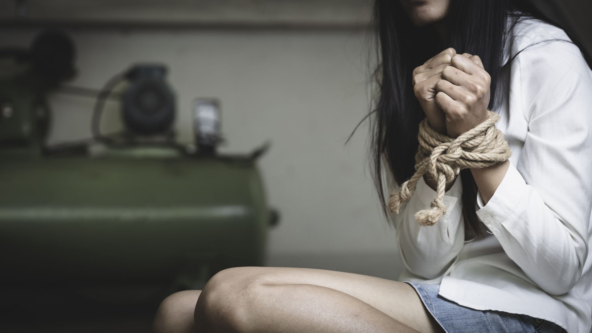 Woman with hands bound by rope, sitting. Blurred background includes industrial equipment.