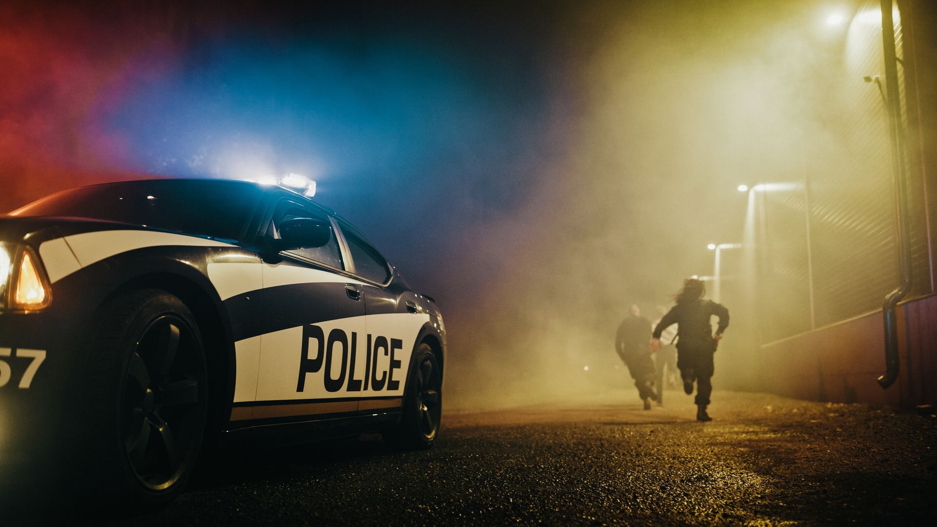 Police car with flashing lights chasing figures running on a wet, foggy street.
