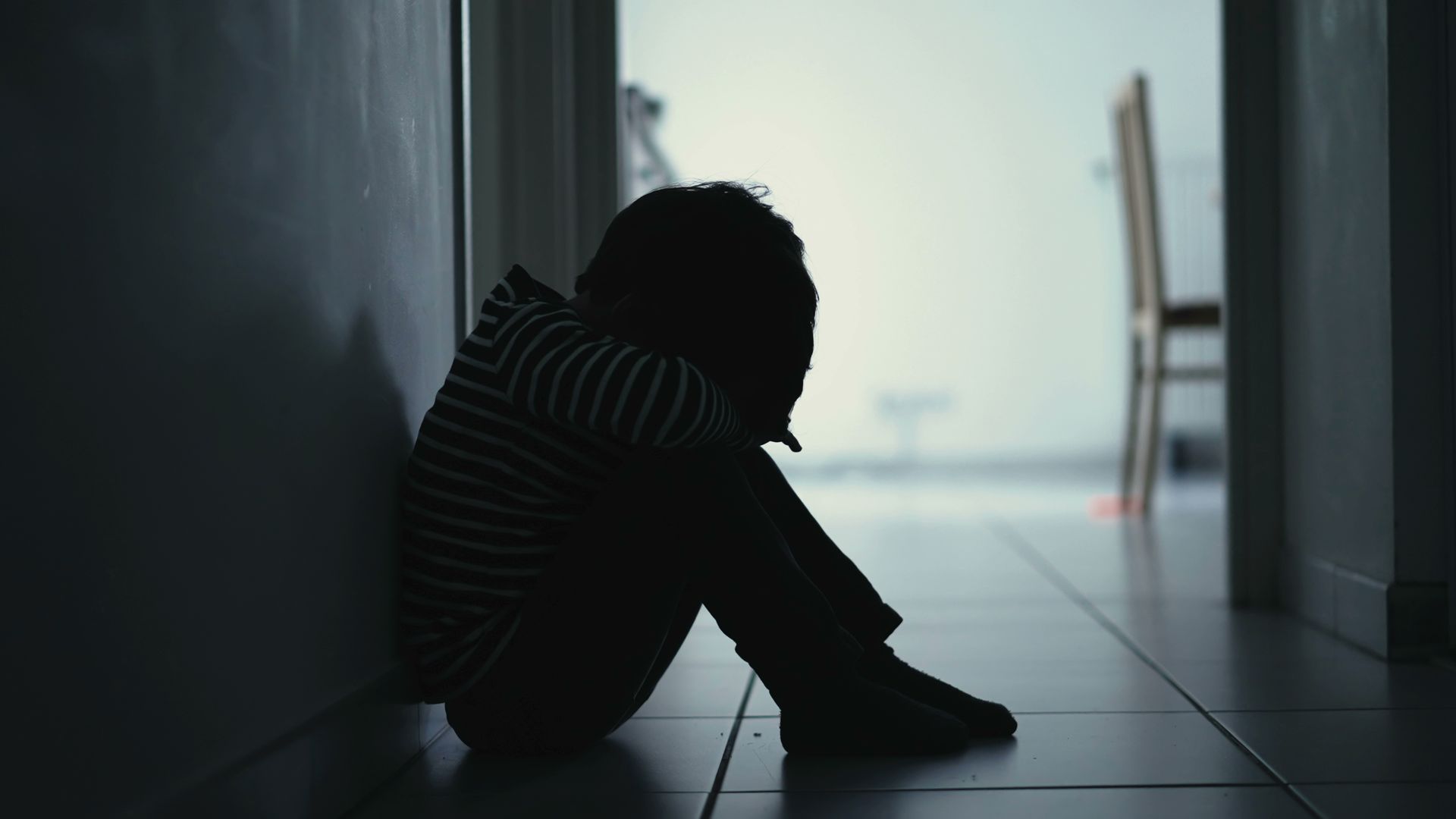 Silhouette of a child, huddled and alone in a hallway, leaning against a wall, appearing distressed.